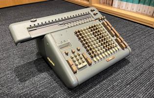 A metal calculator about 24 inches long, 16” wide with number and other control push buttons
